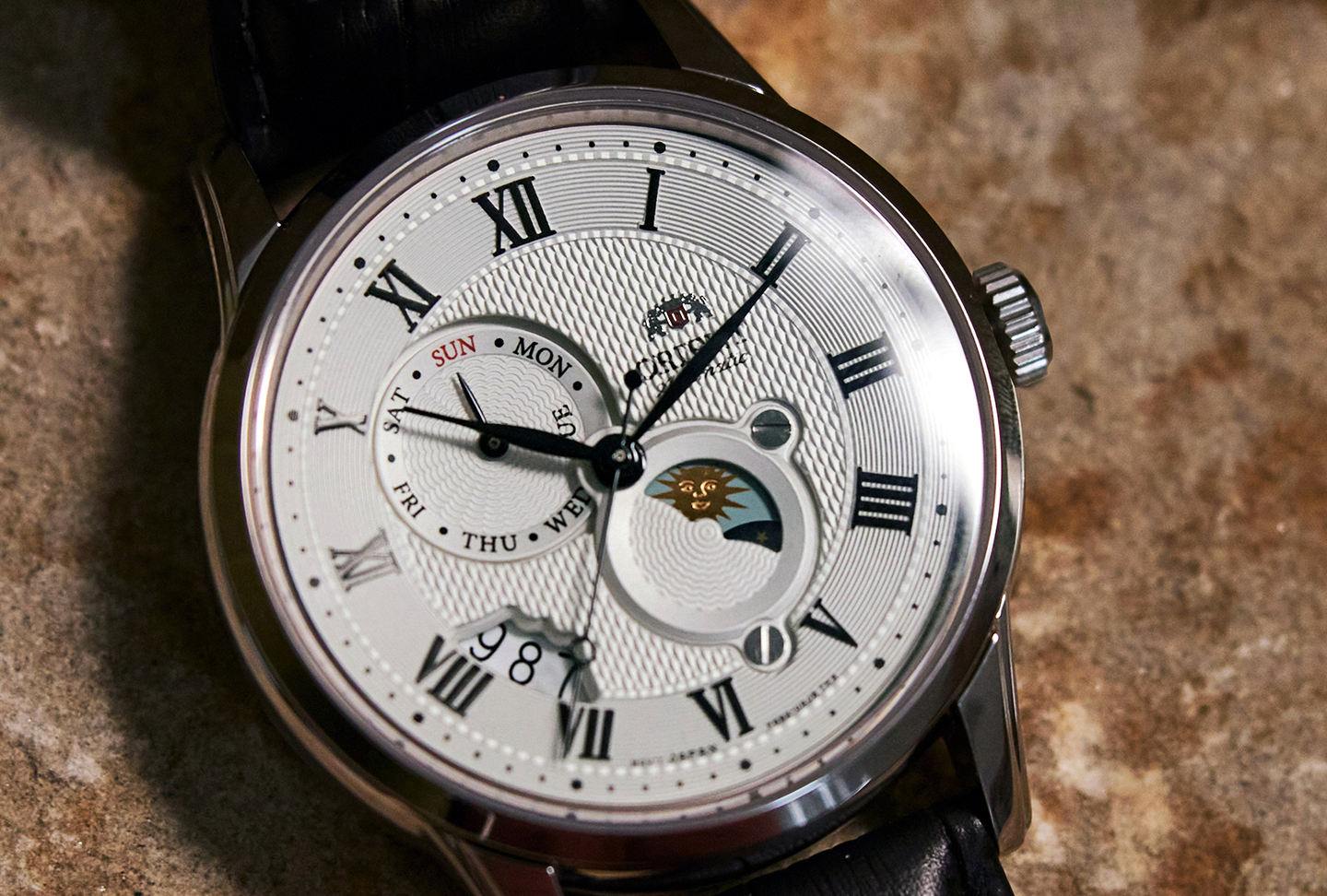 Elegant wristwatch with white textured dial, sapphire crystal, Roman numerals, day and date subdials, and a moon phase display. Black leather strap, classic design.