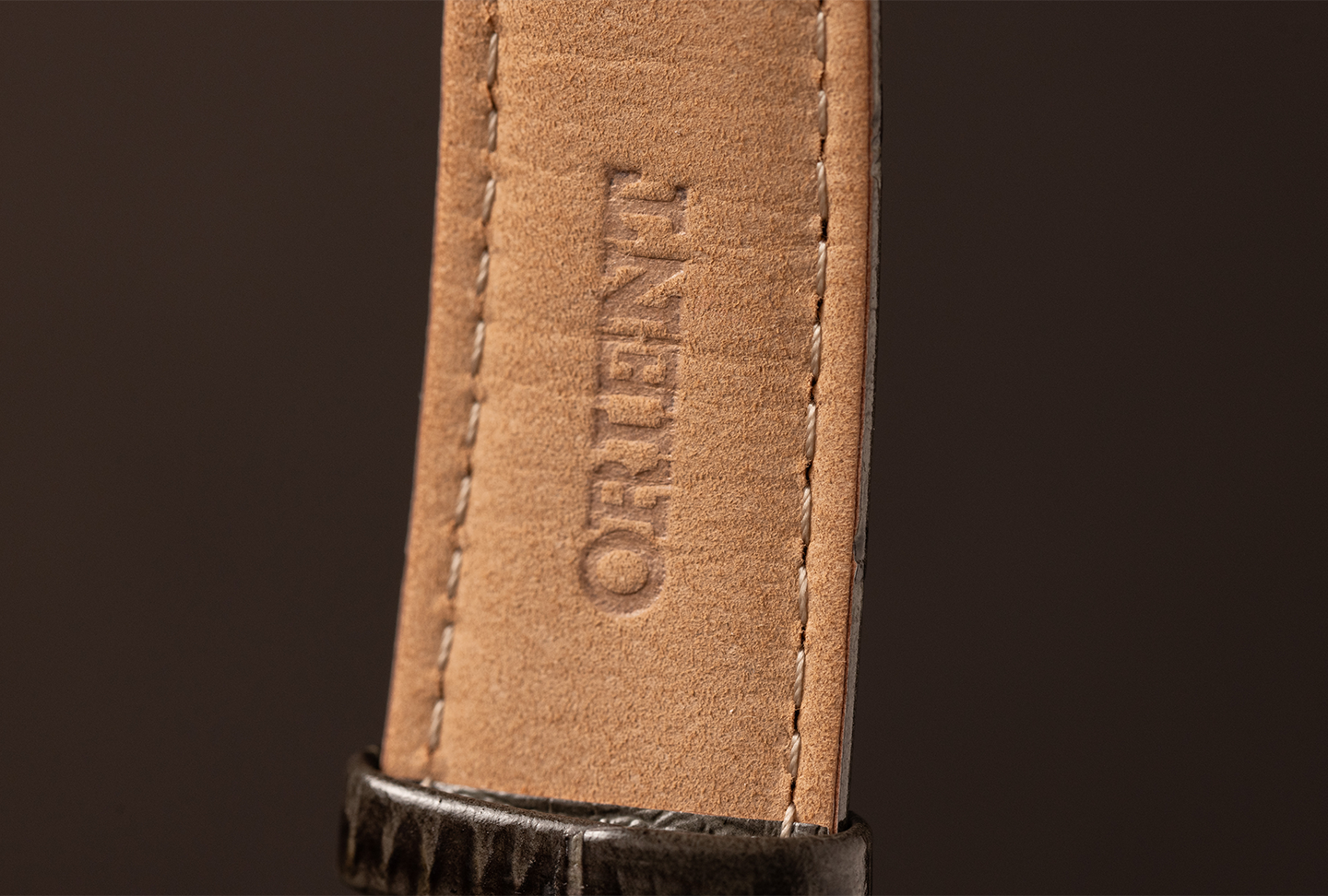 Close-up of a tan leather watch strap with visible stitching, embossed with "ORIENT." The strap's texture contrasts with its dark buckle.