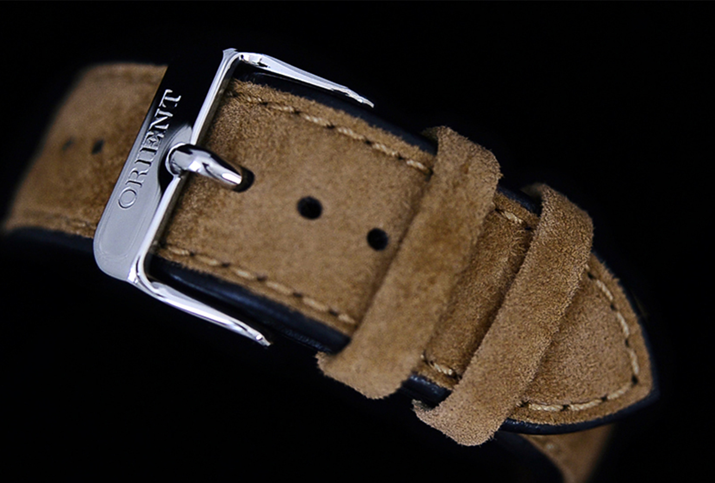 A close-up of a brown suede watch strap featuring a silver buckle with "Orient" engraved. The strap has visible stitching and two loops, set against a black background.