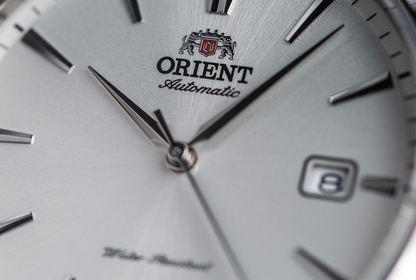 Close-up of an Orient watch face, featuring sleek silver hands and markers, the brand logo at the top, and a date display set to 8. Elegant and refined design.